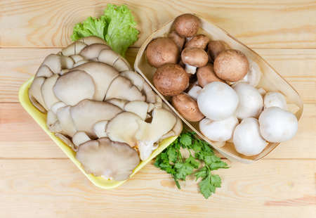 Top view of the cultivated raw brown and white champignon mushrooms in wooden basket, oyster mushrooms on foam food tray and greens on a light colored wooden rustic tableの写真素材