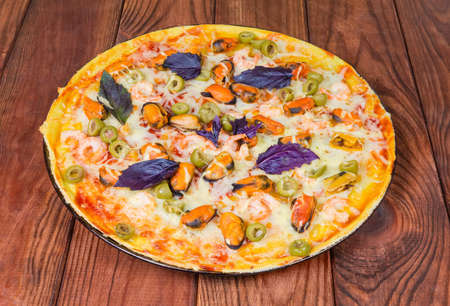 Cooked round pizza with seafoods, olives, decorated with basil on a baking tray on a dark colored rustic wooden tableの写真素材