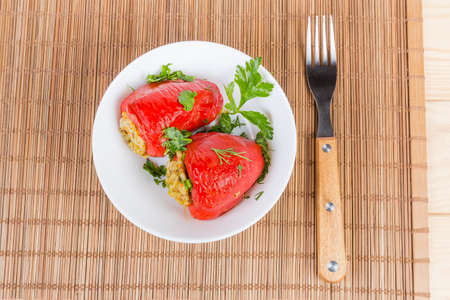 Top view of the two cooked stuffed red bell peppers sprinkled with chopped greens in white bowl and fork on the bamboo table matの写真素材