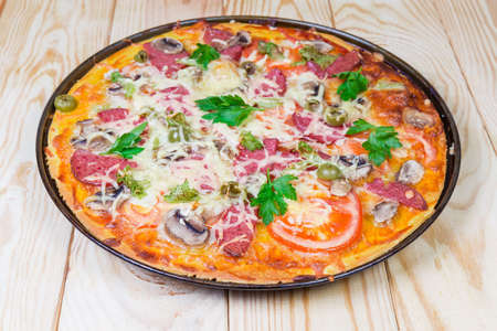 Cooked round pizza with salami, mushrooms, olives and decorated with parsley on a baking tray close-up on a light colored rustic wooden table at selective focusの写真素材