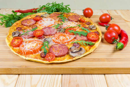 Cooked whole round pizza with different sausages, tomatoes and mushrooms among of greens and vegetables on a bamboo wooden cutting board close-up at selective focusの写真素材