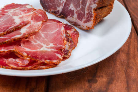 Fragment of white dish with slices and rest of the piece of dried pork neck on a dark colored wooden rustic table close-up at selective focusの写真素材