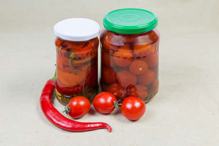 Canned red peppers chili and cherry tomatoes in two different closed glass jars and fresh chili and cherry tomatoes on a fabric surfaceの写真素材