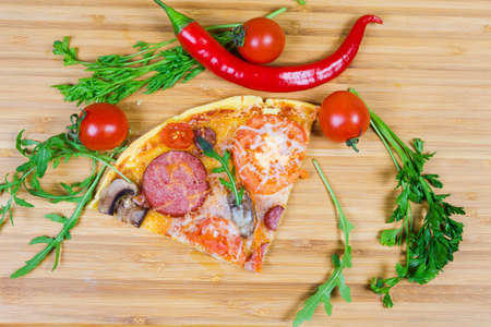Top view of slice of cooked round pizza with different sausages, tomatoes and mushrooms on a bamboo wooden cutting board among of greens and vegetablesの写真素材