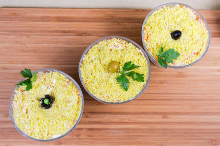 Top view of fish salad with cheese, boiled eggs and vegetables, sprinkled with crumbled egg yolk called as mimosa salad in glass salad bowls on a wooden surfaceの写真素材
