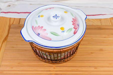 Vintage ceramic soup tureen with closed lid in special thermal insulation wicker wooden basket on a wooden surfaceの写真素材