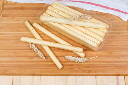 Top view of breadsticks with sesame seeds also known as grissini in transparent plastic container and beside to him, wheat ears on a wooden surfaceの写真素材