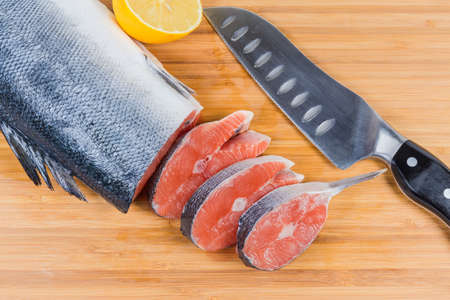 Top view of fresh uncooked salmon steaks among of not sliced piece of fish, lemon half and kitchen knife on a bamboo cutting boardの写真素材