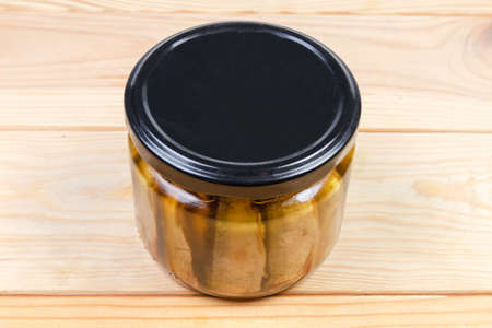 Smoked sprats preserved in vegetable oil in small glass jar with closed black lid on a wooden tableの写真素材