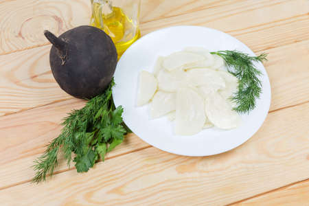 Fresh sliced black radish with salad dressing of vegetable oil and salt on white dish, bottle of oil, greens and whole black radish on wooden rustic tableの写真素材