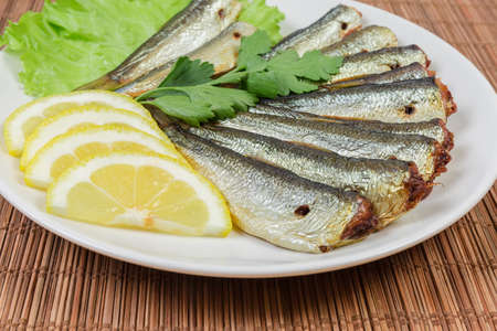 Smoked headless sprats decorated with parsley twig on lettuce leaves, lemon slices on a white saucer...