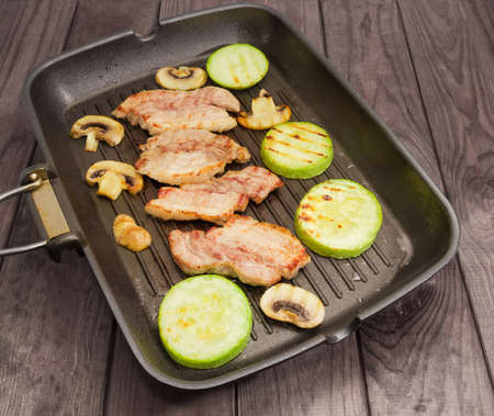 Grilled slices of the streaky bacon, vegetable marrows and mushrooms on the grill pan on the old wooden rustic tableの写真素材