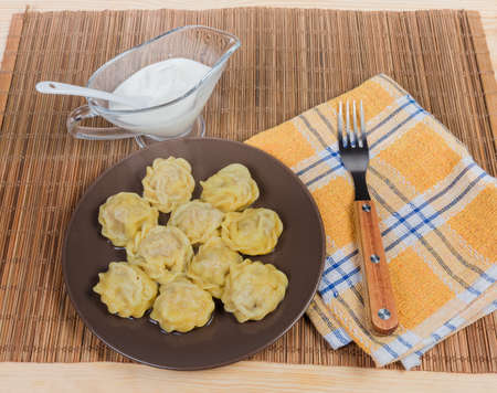 Top view of the cooked homemade meat dumplings, also known as pelmeni on brown dish and sour cream sauce in glass gravy boat, fork on the bamboo table matの写真素材