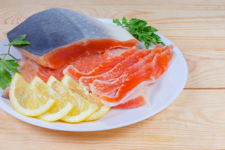Pieces of arctic char fillet on skin and several slices same fish, slices lemon and parslet on white dish on the wooden rustic tableの写真素材