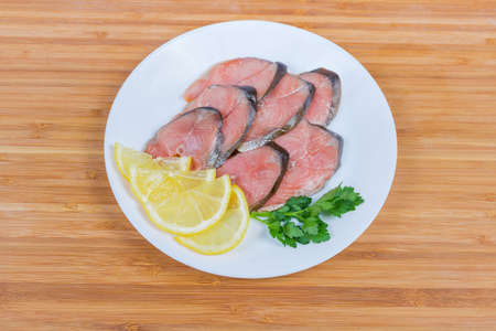 Slices of smoked pink salmon, lemon slices and parsley twig on white dish on the wooden bamboo...