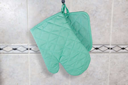 Hanging on a hook set of blue quilted textile oven glove and square pot-holder on a blurred background of the wall covered with gray tileの写真素材