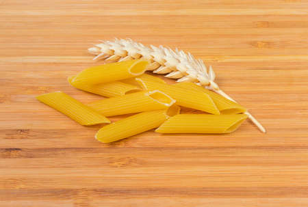 Several uncooked cylinder-shaped grooved pasta also known as penne and ear of wheat close-up on a wooden surfaceの写真素材