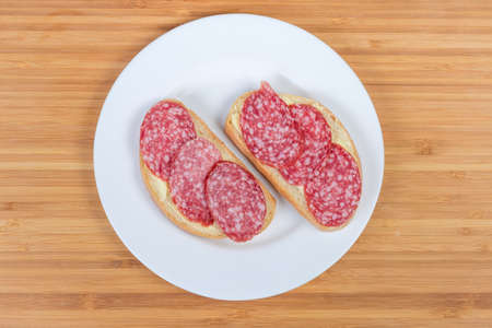 Top view of two open sandwiches made with salami, wheat bread and butter on white dish on a bamboo wooden surfaceの写真素材
