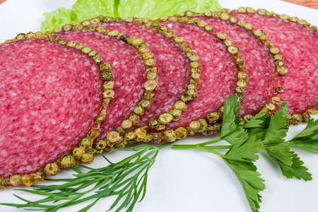 Thin slices of salami, coated with green peppercorns crust, decorated with greens on dish, close-up...