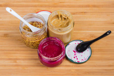 Various condiments - traditional mustard, French whole-grain mustard and horseradish sauce with red beetroot in small glass jars and ceramic spoons on a bamboo wooden surfaceの写真素材