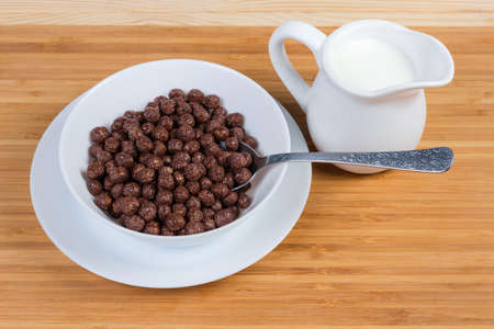 Breakfast cereal chocolate balls in white bowl with spoon, milk in the small milk jug on a wooden surfaceの写真素材