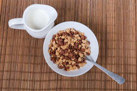 Mix of ordinary and chocolate breakfast cereal with milk addition in the white bowl, milk in the small milk jug, spoon on the bamboo table mat, top viewの写真素材