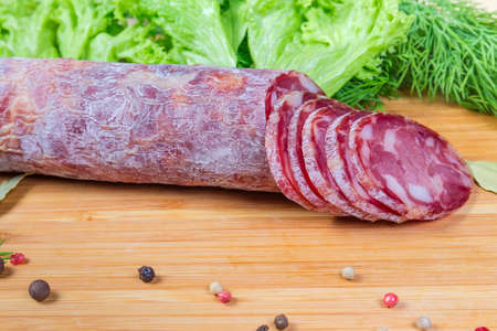 Partly sliced dry-cured sausage among some spices on the wooden cutting board on background of greens, close-up in selective focusの写真素材