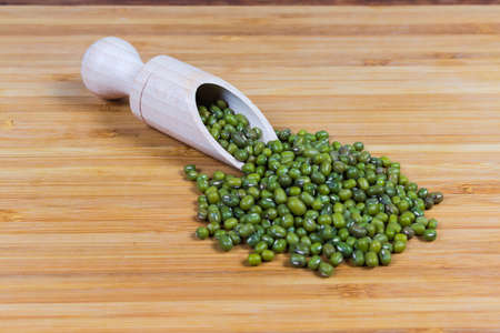 Ripe whole mung beans in wooden spice spoon and scattered beside her on a bamboo wooden surface close-up in selective focusの写真素材