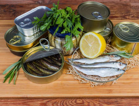 Smoked sprats in cooking oil in the open tin can among of sealed cans and glass jar of canned fish, raw fish greens and lemon on a wooden surfaceの写真素材