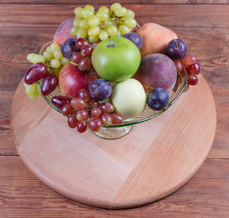 Different fresh fruits in the green glass fruit vase on the round wooden serving board on rustic...