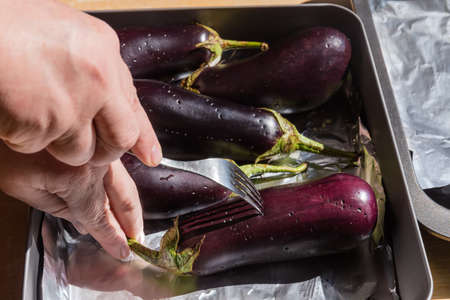 Whole eggplants during puncturing peel with a fork in hand, laid out on the oven tray covered with foil before baking, close-up in selective focusの写真素材
