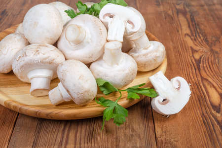 Fresh cultivated common button mushrooms with parsley twigs on the wooden dish on the old rustic table, fragment close-up in selective focusの写真素材
