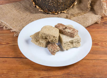 Various crumbly sunflower halva with peanuts, vanilla and cocoa powder addition on white dish on the rustic table on a background of sunflower head with ripe seedsの写真素材