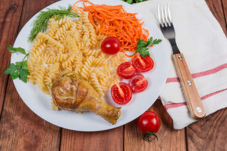 Fried crispy chicken leg with side dish of boiled spiral pasta, carrot salad, fresh cherry tomatoes and greens on the white dish, fork on napkin on the rustic tableの写真素材