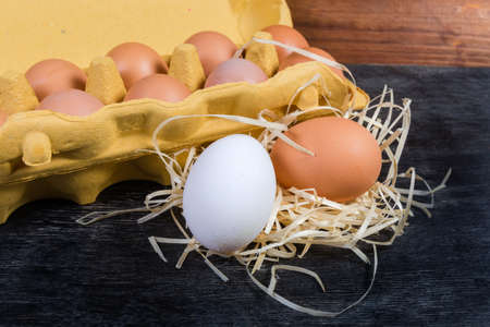 One white and one brown chicken eggs on wood shavings on background of the open egg carton made of...