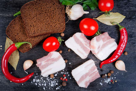 Slices of uncooked pork fatback with layers of lean meat among the some spices and vegetables, brown bread slices on a black surface, top viewの写真素材