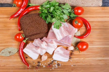 Partly sliced uncooked pork belly with layers of lean meat among the some spices, vegetables and greens, brown bread on the wooden cutting board, top viewの写真素材