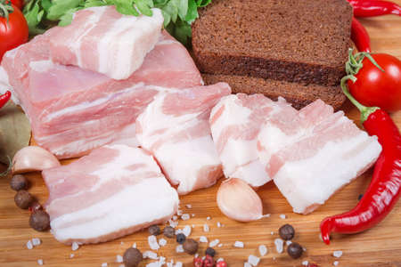 Partly sliced uncooked pork belly with layers of lean meat among the some spices, vegetables and greens, brown bread, close-up on the wooden cutting boardの写真素材