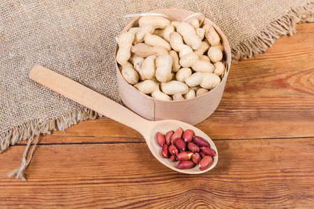 Dried peanuts in their shells in small round wooden box and peeled nuts in the wooden spoon on the...
