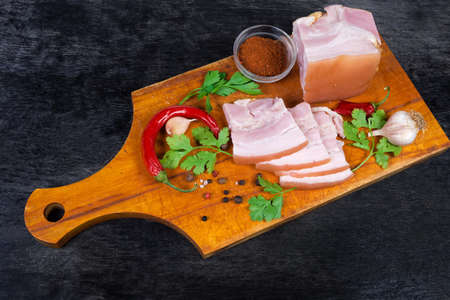 Partly sliced boiled-smoked pork belly among the some spices and greens on the wooden cutting board on the black surfaceの写真素材