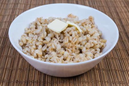 Pearl barley porridge with butter pieces in the white bowl on the bamboo table mat, close-up in selective focusの写真素材