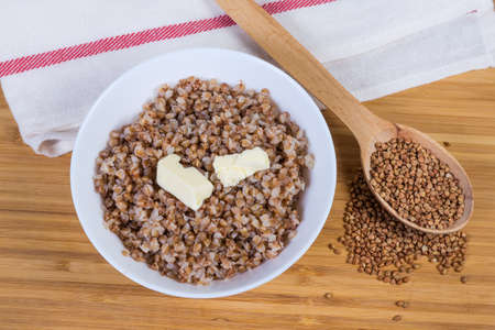 Prepared buckwheat porridge with butter pieces in white bowl and uncooked buckwheat groat in wooden spoon on a wooden surface, top viewの写真素材