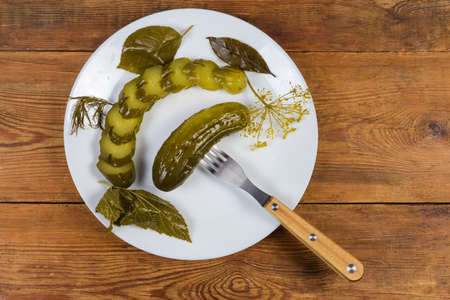 One whole canned cucumber impaled on a fork and sliced cucumber lie among the herbs for canning on the white dish on the rustic tableの写真素材