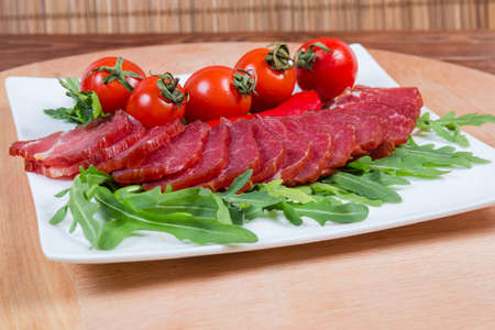 Slices of dried-smoked pork neck among the fresh  greens and cherry tomatoes on dish on wooden surface, close-up in selective focusの写真素材