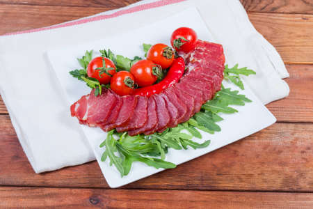 Slices of dried-smoked pork neck among the fresh herbs and cherry tomatoes on the square shape white dish on napkin on the rustic table
の写真素材