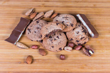 Chocolate chip cookies with nut cream filling among the pieces of milk chocolate and various nuts on the wooden surfaceの写真素材
