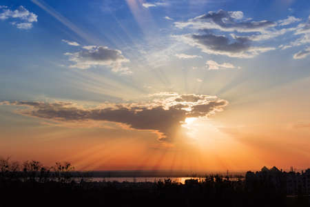 Morning sky with rising sun beams make their way through the clouds over dark suburb and reservoir 