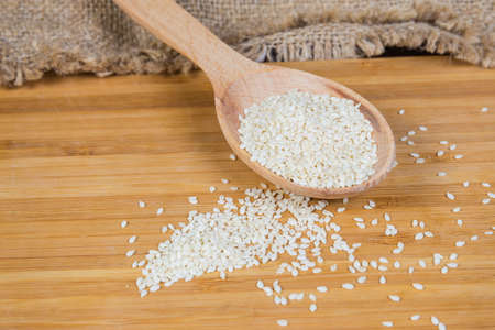 White sesame seeds in the wooden spoon and scattered beside on the wooden surface, close-up in selective focusの写真素材