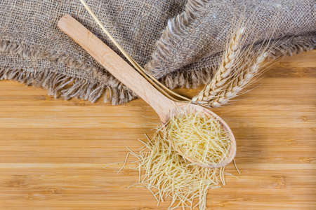 Uncooked very thin short-cut round section pasta known as Filini pasta in the wooden spoon and scatterd beside, wheat ears on wooden surface, top viewの写真素材