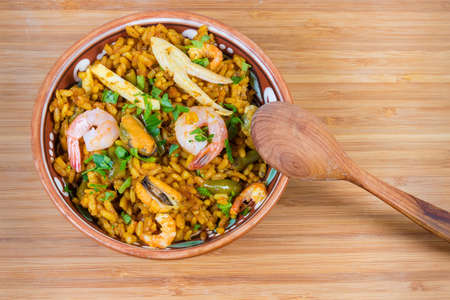 Cooked seafood paella in the clay bowl with wooden spoon on the wooden surface, top viewの写真素材